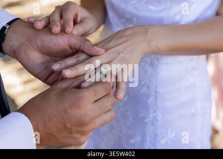 Couple placing halo ring on left finger outdoors in warm sunlight, suit sleeve, watch, white dress Stock Photo