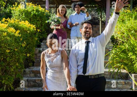 Bride and groom walking down stone steps in garden, bride wearing lace gown, groom waving Stock Photo