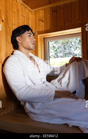 Asian man sitting on wood-paneled sauna bench by small window, wearing white bathrobe with belt Stock Photo