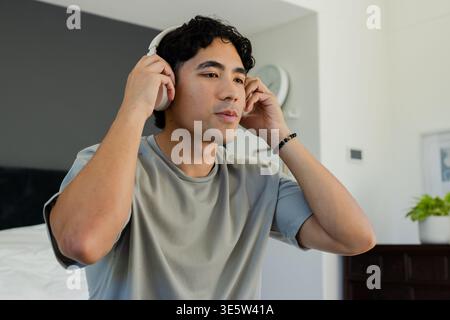 Asian man adjusting white over-ear headphones while wearing grey tee and black bracelet in bedroom Stock Photo