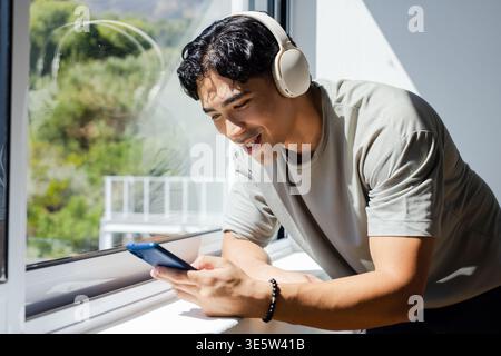 Adult man leaning on windowsill at home smiling checking blue phone with headset and bracelet Stock Photo