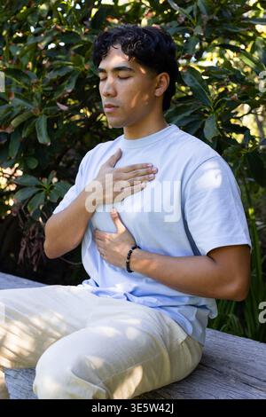 Asian man meditating on wooden bench in park with broadleaf plants, wearing tee  and  beaded bracele Stock Photo