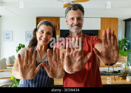 Mature couple standing in open-plan kitchen showing soil-covered palms, wearing striped apron Stock Photo