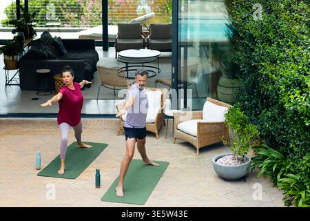 Senior couple doing yoga in activewear on green mats on brick patio near sliding glass doors Stock Photo