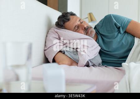 Mature man sleeping on right side in bedroom, wearing teal shirt, pillow and bedside glass visible Stock Photo