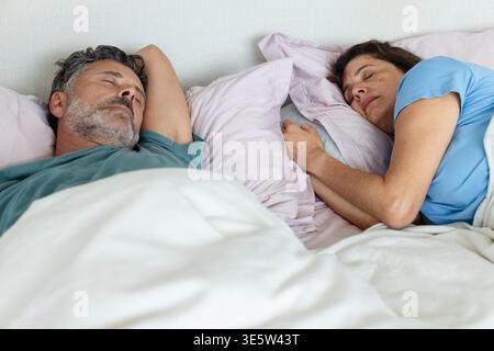 Senior couple lying in bed in bedroom under white duvet, pale pink pillows, wearing short sleeves Stock Photo
