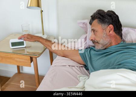 Middle-aged man lying on pink sheets in bedroom reaching toward digital alarm clock in teal shirt Stock Photo