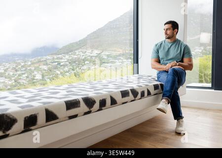 Man sitting on right of window bench in teal T-shirt, wristwatch showing, looking out, copy space Stock Photo