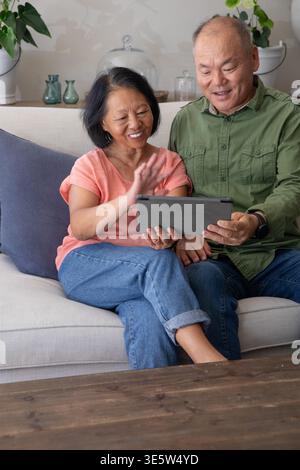 Senior Asian couple sitting on beige sofa sharing tablet, woman coral top man green shirt Stock Photo