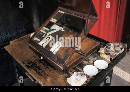Old wooden desk holds vintage photo display box. Glass lid reveals photographs inside. Small white bowls rest on cluttered surface. Various containers Stock Photo