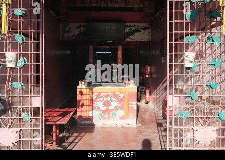 Sunlight streams through an open doorway. Rusty metal grates flank both sides. Green leaves hang from wire frames. Small altar sits at the center. Red Stock Photo