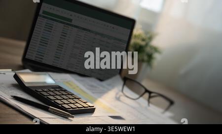 Close-up of a calculator and pen on invoice documents with a laptop displaying a spreadsheet and eyeglasses on a desk, suitable for invoice audit, acc Stock Photo