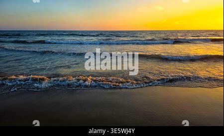 Golden Sunset Waves Rolling onto Sandy Beach. Golden sunset light reflects on gentle waves and wet sand, creating a calm seascape along the shoreline Stock Photo