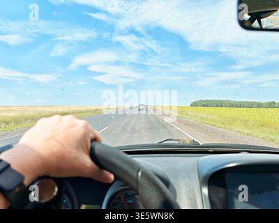 A car drives on an open road with fields on both sides under cloudy skies during autumn. Hands grip the steering wheel. Stock Photo