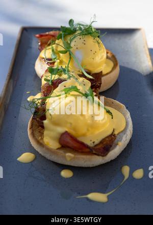 Presented on a slate-gray ceramic tray, these exquisite Eggs Benedict feature golden-toasted English muffins crowned with crisp, salt-cured bacon and Stock Photo