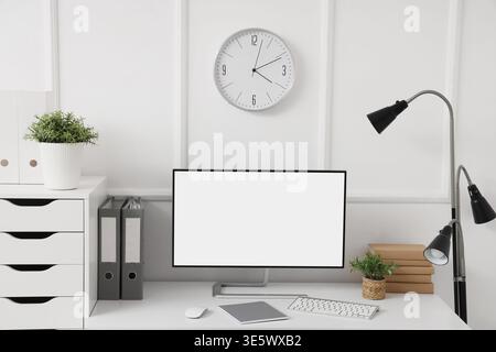 Workplace with blank computer monitor, lamp and clock on light wall in office Stock Photo