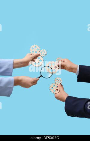 Hands with wooden gears and magnifier on blue background Stock Photo