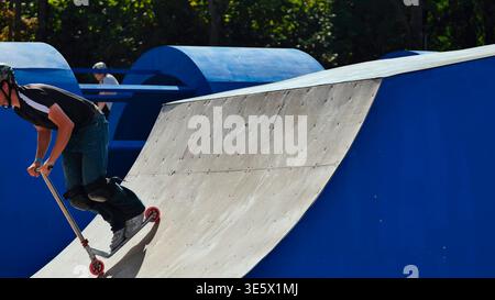 Young man riding a scooter on a ramp at an outdoor skatepark, performing tricks Stock Photo