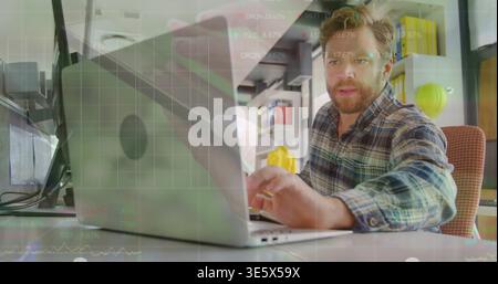 Working bearded man wearing plaid shirt typing on laptop in small office, with yellow hard hat Stock Photo