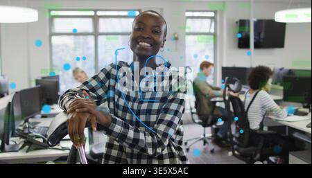 Smiling woman wearing checkered shirt leaning on chair in open office, with monitors, blue overlay Stock Photo