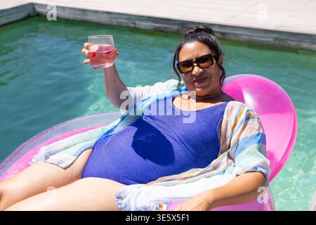 Sun urging African woman reclining on pink float in pool, wearing blue swimsuit, holding glass Stock Photo