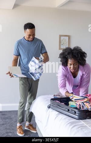 African American couple packing black hard-shell suitcase on bed, man holding notebook, copy space Stock Photo