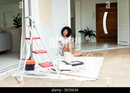 African American woman sitting in entry using phone in overalls amid ladder, paint cans, copy space Stock Photo