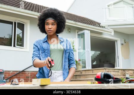 African woman wearing denim shirt at porch workbench measuring wooden plank with tape measure Stock Photo