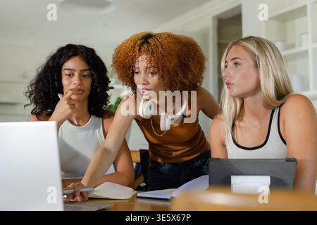Three female friends collaborating around table in kitchen, pointing at laptop wearing headphones Stock Photo