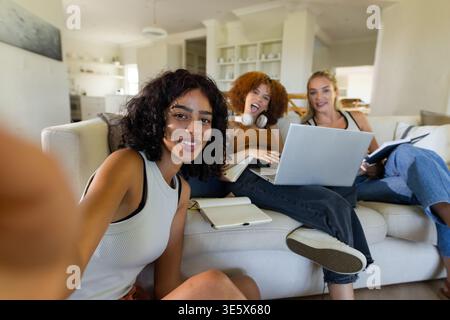 Female friends sitting on light sofa at home taking selfie with smartphone, using laptop, notebooks Stock Photo