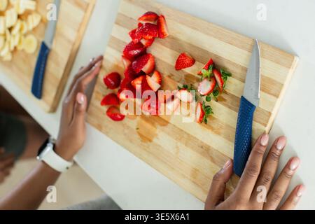 Two female friends slicing strawberries on kitchen counter using wooden cutting board and knife Stock Photo