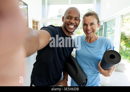 Couple in athletic shirts posing while taking phone selfie at home holding yoga mats water bottle Stock Photo