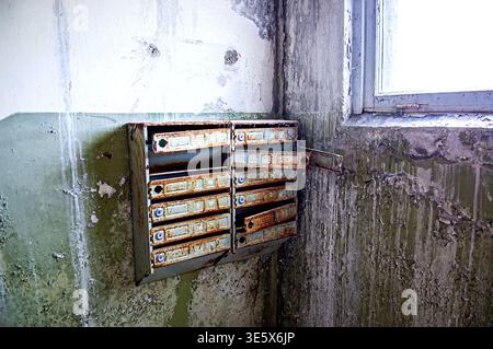 Ukraine, Pripyat : Set of old metal mailboxes heavily rusted and open mounted on peeling painted wall next to window in the entrance hall of an abando Stock Photo