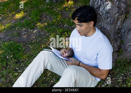 Asian man sitting on grass by trunk writing journal with pencil white-tee beige-pants, copy space Stock Photo