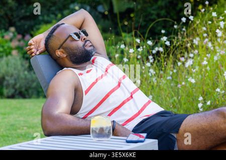 Mid-adult African man reclining on chair on mown lawn in tank top, glass and smartphone nearby Stock Photo