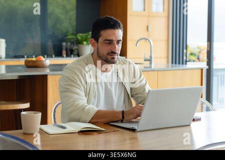 Man working on silver laptop at wooden dining table in modern kitchen, notebook, ceramic mug nearby Stock Photo