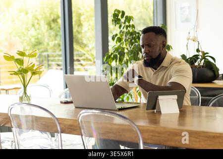 Adult African man checking smartwatch while working on silver laptop at home with tablet smartphone Stock Photo