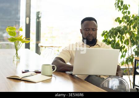 Man working on silver laptop at wooden table by glass windows with green mug and notebook Stock Photo