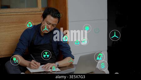 Writing man in black apron at storefront counter, using notebook pen by open laptop and icons Stock Photo
