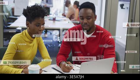 Working at office desk, woman in yellow top, man in red sweater reviewing laptop papers, overlays Stock Photo