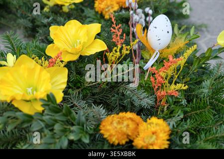 Decorative Easter arrangement with yellow flowers and a painted egg ornament is placed among evergreen branches in an outdoor market setting Stock Photo