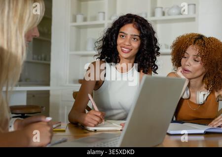 Diverse female friends studying together at home kitchen table with silver laptop and notebooks Stock Photo