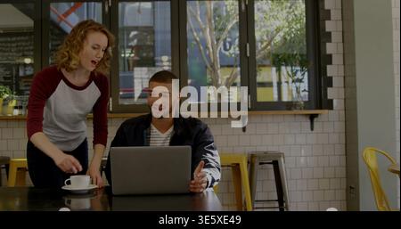 Working man using silver laptop at cafe, woman in burgundy sleeves leaning in, holding coffee cup Stock Photo