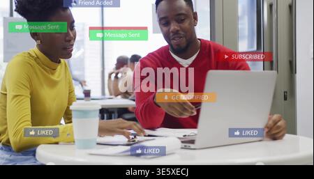 Pointing man in red sweater guiding woman yellow top at shared workspace, laptop and badges showing Stock Photo
