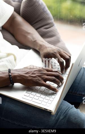 Adult man typing on white laptop at home on sofa cushion, wearing blue jeans, beaded bracelet Stock Photo