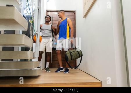 Standing diverse male friends in home foyer, holding orange basketball and green duffel bag Stock Photo