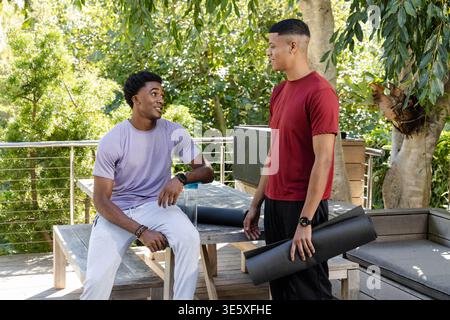 Diverse male friends sitting and standing on backyard deck, with exercise mat and water bottle Stock Photo