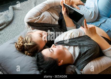 Happy two sisters using tablet while lying on their backs on floor at home laughing cheerfully watching videos or chatting online, wearing gray clothes Stock Photo