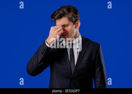30s aged businessman in formal wear having headache, studio portrait. Guy putting hands on head, isolated on blue background. Concept of problems, medicine, illness Stock Photo
