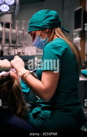 Female dentist performing dental treatment on adult patient in modern dental clinic wearing scrubs and mask Stock Photo